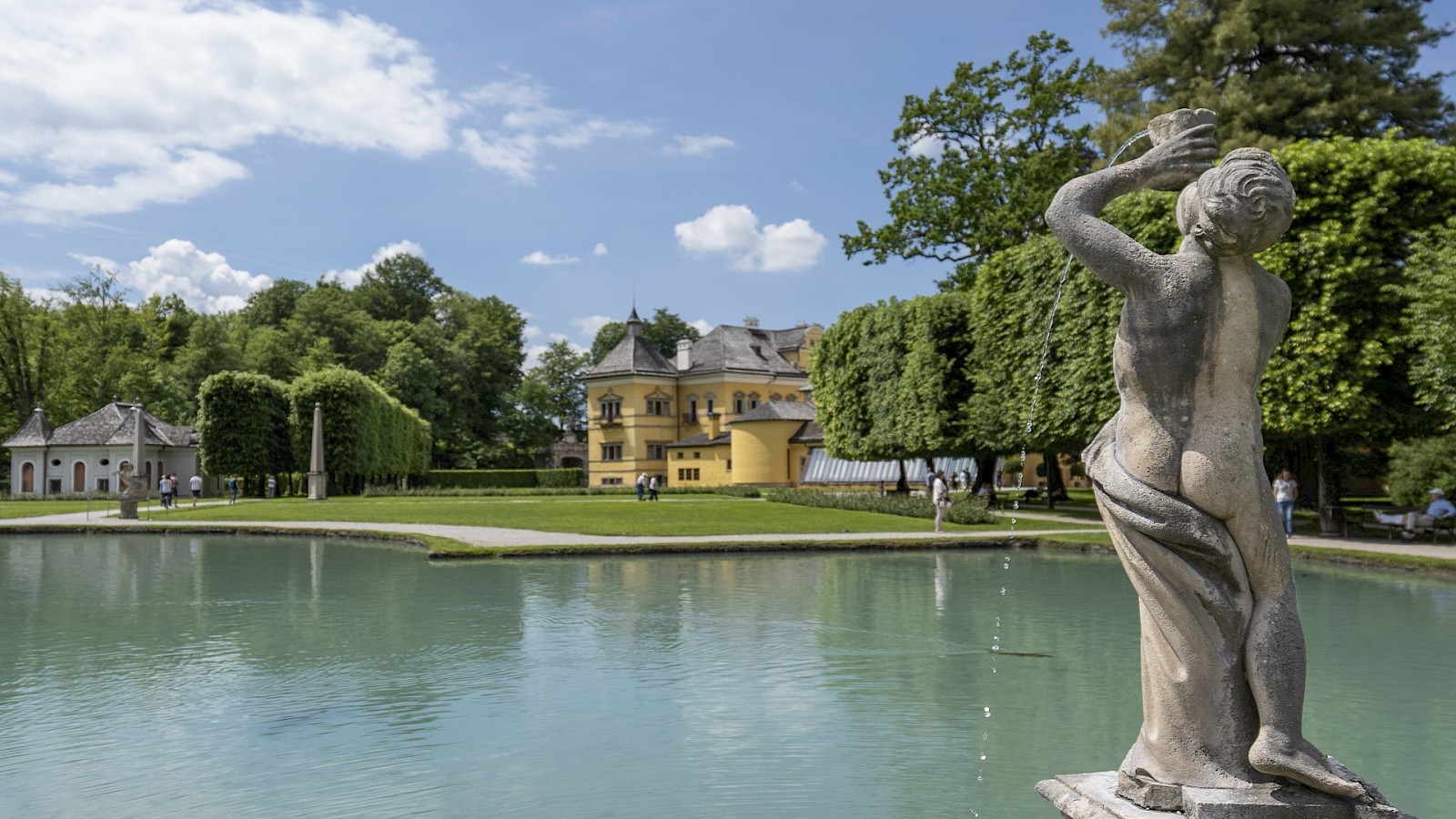 Schlosspark Hellbrunn copyright Gassner Gastronomie:  Das Bild zeigt eine malerische Parklandschaft mit einem barocken Schloss im Hintergrund, vermutlich das Schloss Hellbrunn in Salzburg. Im Vordergrund befindet sich eine steinerne Brunnenfigur, aus deren erhobener Muschel Wasser in einen ruhigen Teich fließt. Umgeben ist die Szene von gepflegtem Rasen, klar geschnittenen Hecken und hohen Bäumen, die Schatten spenden. Einige Besucher spazieren durch die Anlage oder genießen die Aussicht. Der Himmel ist blau mit vereinzelten weißen Wolken, was der Szene eine friedliche und sommerliche Atmosphäre verleiht. Die Harmonie zwischen Natur und Architektur ist besonders beeindruckend.