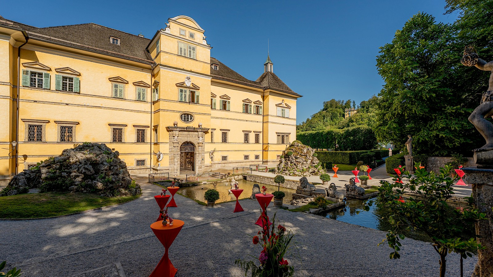 Schlosspark Hellbrunn copyright Gassner Gastronomie:  The image shows a picturesque park landscape with a Baroque-style palace in the background, likely Hellbrunn Palace in Salzburg. In the foreground, there is a stone fountain statue pouring water from a raised shell into a serene pond. The area is surrounded by well-maintained lawns, neatly trimmed hedges, and tall trees providing shade. A few visitors are walking through the grounds or enjoying the scenery. The sky is blue with scattered white clouds, adding to the peaceful, summery atmosphere. The harmony between nature and architecture creates an elegant and tranquil setting.