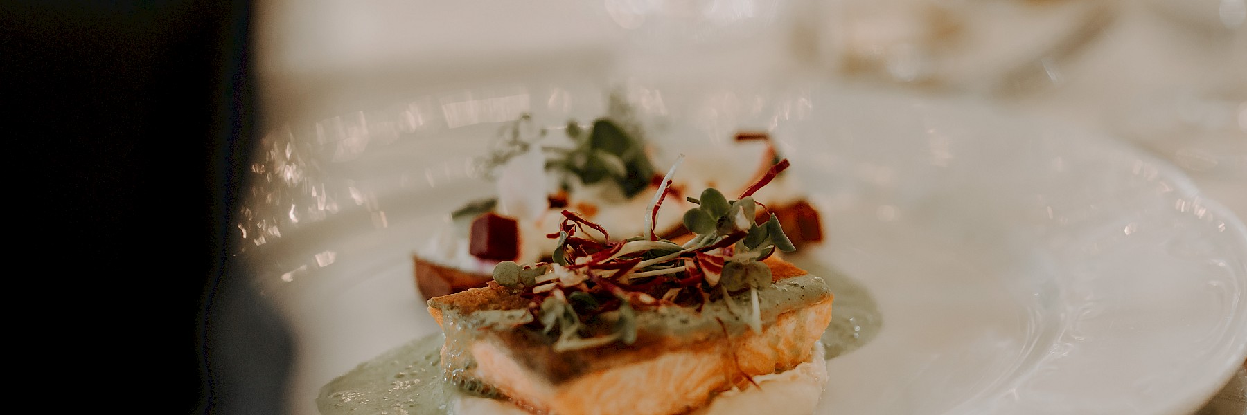 Schlossrestaurant Hellbrunn (c) Gassner Gastronomie, Fotografie Farbenspiel:  The image shows an elegantly plated gourmet dish. At the center is a perfectly cooked piece of salmon, resting on a creamy purée and topped with a light green sauce and fresh microgreens. Behind the salmon are additional elements, possibly roasted beet cubes and a dollop of herbed cream or cheese, artfully arranged. The plate is fine white porcelain, complemented by polished cutlery and a softly lit background with glassware, suggesting a formal dining setting. The dish is visually appealing and clearly crafted with attention to detail—ideal for a refined dinner occasion.