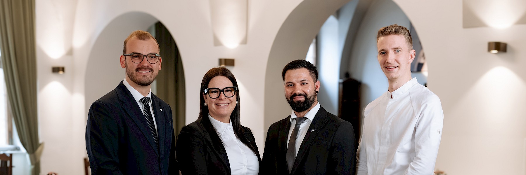 Team photo (c) Gassner Gastronomie, Foto: Hechtfisch: In the image, you see a friendly team standing together in a bright, elegant room. Four staff members stand side by side, smiling warmly. Two people wear formal suits, one person wears a black business outfit with a white blouse, and one person is dressed in a light-colored chef’s jacket. In the background, there are large arches, soft lighting, and tasteful decorations. The space feels calm, clean, and high-quality. The atmosphere is welcoming and professional. The team gives a sense of competence and warmth. Guests can feel comfortable here and expect a pleasant stay or a special dining experience.