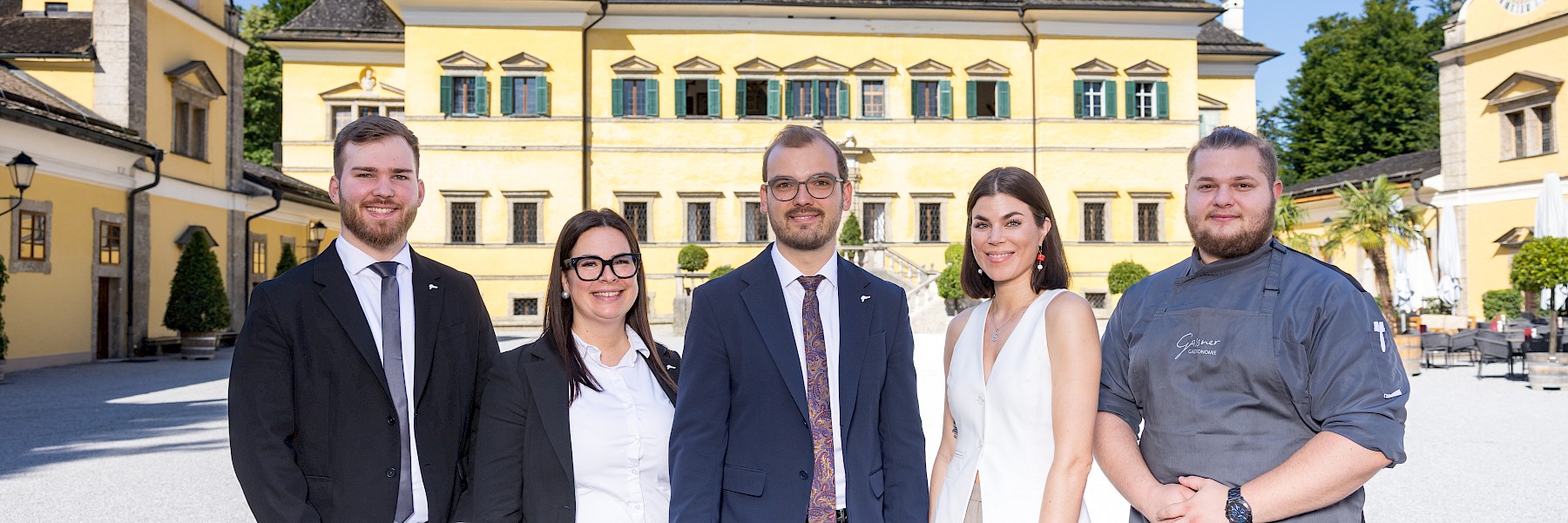 In front of the historic backdrop of Schloss Hellbrunn, part of the Gasthaus team stands together in a professional and elegant appearance. Five team members are side by side – three in stylish suits, one in light business attire, and one in a modern chef’s uniform. They radiate warmth, confidence, and strong team spirit. The yellow façade of the castle and the clear blue sky create a welcoming and vibrant atmosphere. This image perfectly captures the blend of tradition, hospitality, and contemporary culinary culture that defines the essence of the Gasthaus zu Schloss Hellbrunn.
