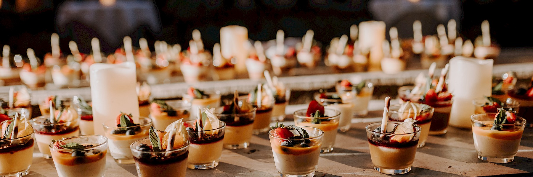 Dessertbuffet Hellbrunn (c) Gassner Gastronomie, Fotografie Farbenspiel: The image shows a beautifully arranged dessert station at dusk. Numerous small glasses filled with artfully prepared desserts—garnished with berries, mint, cream, and other delicate toppings—are neatly lined up on a table. Soft, warm lighting creates an elegant and inviting atmosphere. White candles placed between the glasses add to the ambiance, casting a gentle glow across the scene. The setting evokes a high-end event or wedding celebration, radiating attention to detail and culinary finesse. It’s a feast for the eyes and a promise of delicious indulgence.