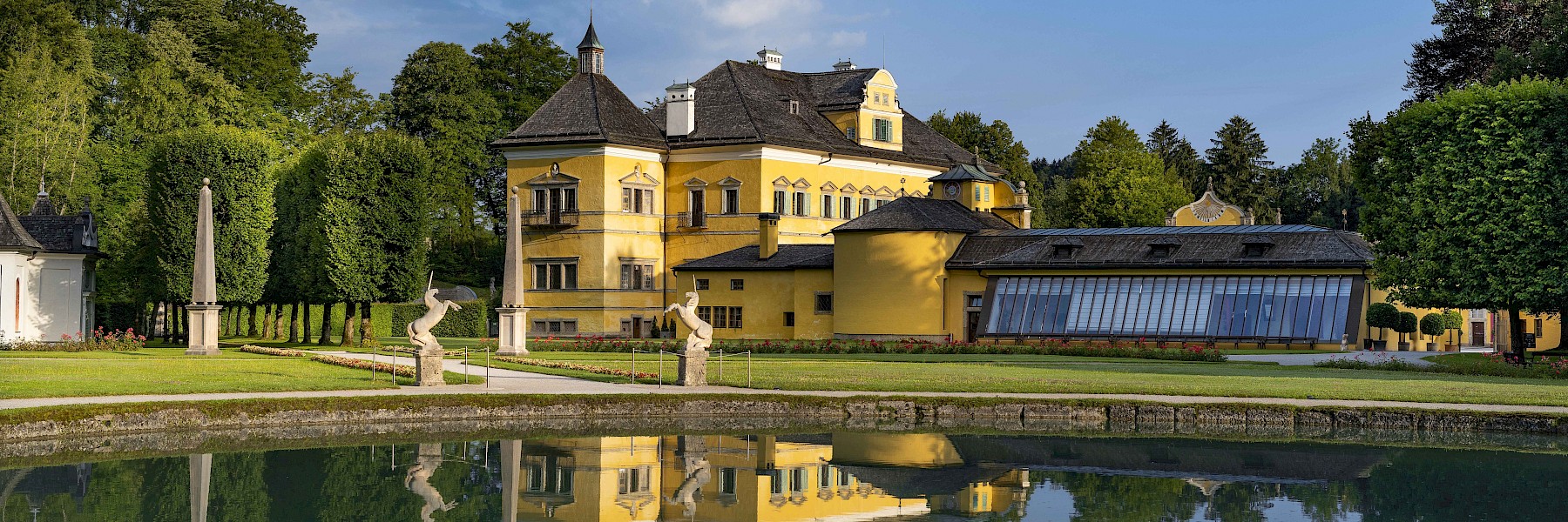 Hellbrunn Schloss Wasserparterre (c) Schlossverwaltung Hellbrunn Foto Auer: The image shows Hellbrunn Palace in Salzburg on a sunny day, beautifully nestled within a manicured park landscape. The baroque architecture of the yellow palace is clearly reflected in the calm waters of the pond in the foreground. Surrounded by lush greenery, neatly trimmed hedges, and elegant sculptures, the scene conveys a sense of tranquility and grandeur. Notable features include the decorative façade details and the distinctive roof with small towers. This setting radiates historical elegance and timeless charm—a place where history, art, and nature blend in perfect harmony.