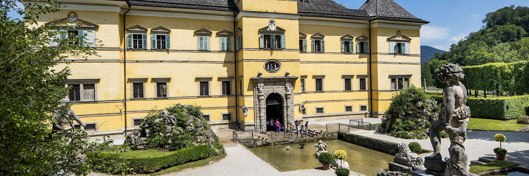 Hellbrunn Schloss (c) Schlossverwaltung Hellbrunn Foto Auer:  The image shows the impressive façade of Hellbrunn Palace in Salzburg, a Baroque pleasure palace painted in a warm yellow with green shutters. In front of the grand entrance lies a neatly maintained courtyard with a rectangular reflecting pool, framed by stone statues and symmetrically arranged plants. A group of visitors gathers at the entrance, adding life to the serene setting. Surrounding the palace are manicured hedges, lush lawns, and ornamental garden features. In the background, forested hills rise under a clear blue sky, creating a picturesque and elegant scene rich in historical charm.