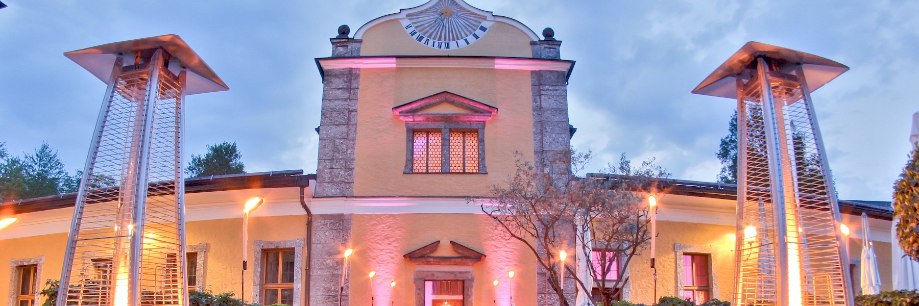 Gasthaus Hellbrunn (c) Gassner Gastronomie: The image shows the festively illuminated entrance of the guesthouse at Hellbrunn Palace, with a red carpet leading to the main entrance. Modern patio heaters and torches line both sides of the path, creating an elegant and warm atmosphere. The building’s façade is painted a soft yellow and features stone accents, along with a striking gable that holds a sundial. In front of the entrance, white-clothed tables are set up – suggesting an upcoming event or an exclusive dinner.