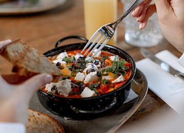 Schlossrestaurant Hellbrunn (c) Gassner Gastronomie: Das Bild zeigt eine köstliche Brunchkreation im Schlossrestaurant Hellbrunn. Im Fokus steht ein frischer Sesam-Bagel, liebevoll belegt mit Avocadoscheiben, zartem Räucherlachs, cremigem Frischkäse und einem pochierten Ei, garniert mit frischen Microgreens. Das Gericht wird auf einem stilvollen Keramikteller serviert, begleitet von einem Glas Sekt, was dem Moment eine festliche Note verleiht. Das warme Sonnenlicht und das rustikale Holzambiente schaffen eine einladende, entspannte Atmosphäre – perfekt für einen genussvollen Start in den Tag.