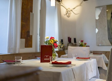 The image shows a stylishly set table at Schlossrestaurant Hellbrunn. A white tablecloth and red napkins create an elegant and inviting atmosphere. A small bouquet of colorful flowers adds a fresh, cheerful accent to the table. In the center stands a dark red menu with the restaurant’s logo. The room is bright and welcoming, enhanced by soft daylight filtering through white curtains. The interior blends rustic charm—like the mounted antlers on the wall—with modern art, creating a harmonious setting that is perfect for a refined dining experience.