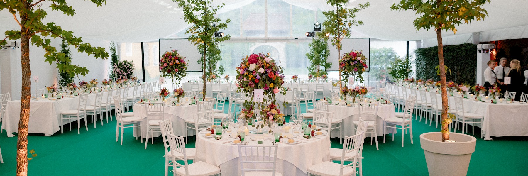Hellbrunner Adventzauber (c) Gassner Gastronomie, Foto Zauner: The image shows an elegant event tent beautifully decorated for a wedding or special celebration. White round tables are set with fine tableware, white chairs, and vibrant floral centerpieces, creating a festive and refined atmosphere. Tall flower arrangements and potted green trees add a fresh, natural touch to the space. The green carpet complements the botanical theme, adding a lively feel. Soft daylight filters through the transparent tent walls, giving the setting a bright and welcoming ambiance—perfect for a stylish summer celebration or garden-inspired event.