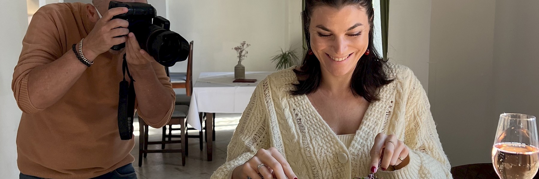 Hellbrunn Schloss Wasserparterre (c) Schlossverwaltung Hellbrunn Foto Auer: The image shows a young woman in a stylish, light-filled room enjoying a beautifully presented burger. She smiles as she cuts into the dish with a knife and fork. To her right stands a glass of rosé sparkling wine. On the left side of the image, a photographer with a professional camera captures the moment with focused attention. The scene feels natural and lively—a blend of culinary enjoyment and professional food photography. The soft lighting and elegant setting enhance the refined and inviting atmosphere of this moment.